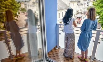 Two women are standing on a balcony, looking at a lively street. The view shows shops and people, while the women’s reflections can be seen in the glass.