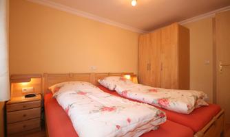 A cozy bedroom with two single beds and light walls. The room is simply furnished with a wooden wardrobe and night lamps.