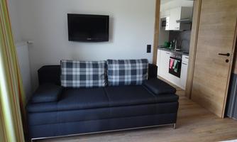 A cozy living room with a black sofa and a television on the wall. In the background, a modern kitchen is visible.