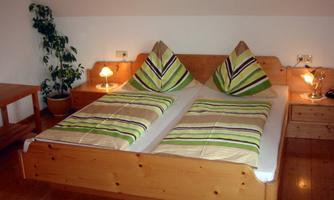 A cozy bedroom with a double bed and colorful bedspreads. The furniture is made of wood, and next to the bed are table lamps and a plant.