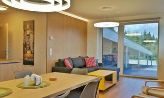 A modern living room with a cozy couch and a dining table. Large windows offer a view outside.