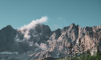 An impressive mountain landscape with high peaks and scattered clouds. In the foreground, there are green coniferous trees.