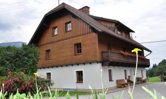 A traditional wooden house with a balcony and blooming flowers in the foreground. In the background, gentle hills and a cloudy sky can be seen.
