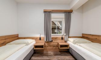 A simple room with two single beds and wooden paneling. The window is equipped with curtains and offers a view outside.