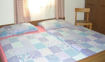 A cozy bedroom with a large double bed and colorful bedspreads. Next to the bed is a chair, and the window lets in soft light.