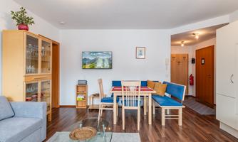 A cozy living space with a dining table and chairs. A landscape painting hangs on the wall, and there is a glass display cabinet as well as a couch.