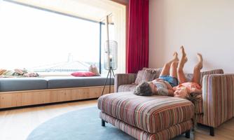 A cozy living room corner with a large window. Two people are relaxing on a sofa and a pouf.