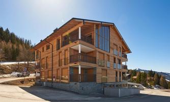 A modern wooden building in a mountainous setting. The facade has large windows and balconies, with a clear blue sky in the background.