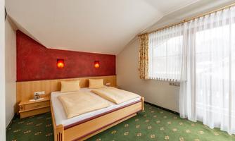 A cozy bedroom with a double bed and cream-colored pillows. Large windows let in plenty of light and the carpet has a blooming pattern.
