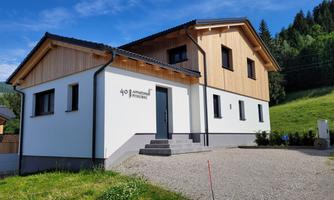 A modern residential building with a wooden and plaster façade, surrounded by green meadows. The architecture combines stylish elements with an inviting entrance.