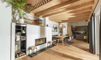 A modern living room with a wooden beam ceiling and a cozy dining table. The space is bright and inviting, with plants and a fireplace as a focal point.