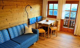 A cozy room with a blue sofa, a wooden table, and chairs. Large windows provide a view outside and let in plenty of light.