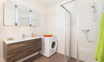 A modern bathroom with a shower and a washing machine. There is a wooden cabinet with a sink and mirrors.