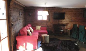 A cozy living room with red couch furniture and rustic wooden walls. A dark carpet is on the floor, and there is a TV as well as a dining table in the background.