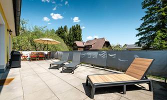 A sunny terrace with loungers and a sunshade. In the background, trees and houses are visible.