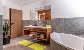 A modern bathroom with a freestanding bathtub and a wooden vanity. The walls are in neutral shades of gray, and there are some green towels and a plant.