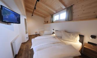 A modern bedroom with a double bed and high wooden ceilings. A television is mounted on the wall and large windows provide plenty of daylight.