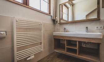 A modern bathroom with a wooden vanity and a large mirror. A radiator is mounted on the wall, and the windows provide plenty of natural light.
