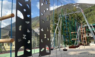 A playground with various play equipment such as swings and climbing elements. In the background, mountains and a blue sky are visible.