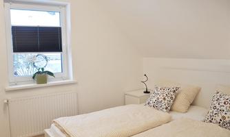A cozy bedroom with a double bed and two cushions. The room has a window with blinds and a small plant on the nightstand.
