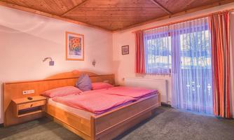 A cozy bedroom with a wooden bed and red bedding. Large windows let in a lot of light.