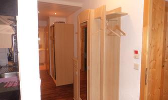 A narrow hallway with wooden cabinets and a mirror. Two coat hangers are hanging on the wall, and the floor is laid with wooden planks.