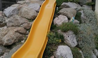 A yellow slide is placed on a slope made of stones and plants. The area is green and surrounded by nature.