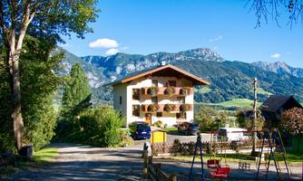 A charming house with balcony flowers, surrounded by green nature and mountains. In the foreground, there is a playground for children.