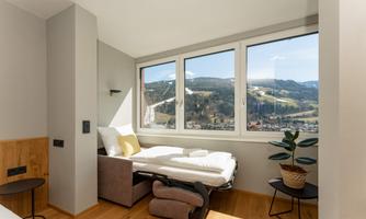 A bright room with a beautiful view of the mountains. There is a bed, a cozy armchair, and a plant.