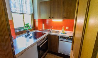 A small kitchen with wooden cabinets and a red tiled wall. The countertop is clean and well-equipped.