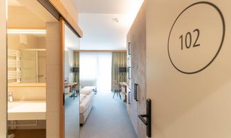 A modern hotel room entrance with room number 102. In the background, a bright room and a bathroom are visible.