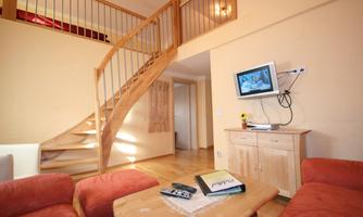 A cozy living space with a wooden staircase and a TV on the wall. The furniture is simple and inviting, with a light color tone.