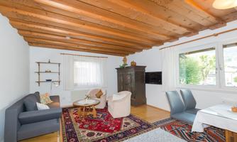 A cozy living room with wooden ceilings, a carpet, and modern furniture. Large windows let in plenty of light and provide a view outside.
