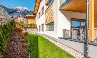 A modern building with wooden cladding and large windows. The well-kept garden with green lawn and gravel areas complements the beautiful view of the mountains.