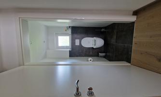 A modern bathroom with a white toilet and dark floor. Through the open door, a window is visible, allowing natural light to enter.