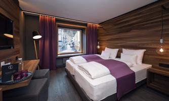 A modern furnished hotel room with a large window overlooking a street. The walls are clad in wood, and the beds are dressed in white linens and purple blankets.