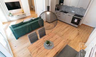 A modern, open plan kitchen with a cozy green sofa and a large wooden table. The room has bright wooden floors and attractive lighting.