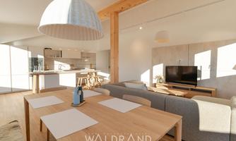 A modern living room with a wooden dining group and an open kitchen. Bright colors and plenty of daylight create an inviting atmosphere.
