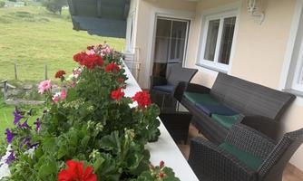 A cozy balcony with garden furniture and colorful flowers. In the background, a green landscape can be seen.
