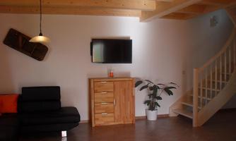 A bright living space with a black couch and a wooden piece of furniture. A staircase leads to the upper area, and a television is hanging on the wall.