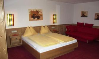 A cozy bedroom with a double bed and yellow pillows. Decorative pictures hang on the wall, and a red sofa sits beside it.