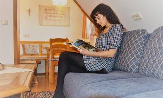 A woman is sitting on a couch and reading a magazine in a cozy room. In the background, there are wooden-paneled walls and a dining table visible.