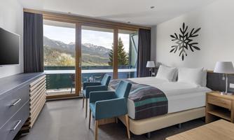 A modern hotel room with large windows and a view of the mountains. The decor is simple and elegant, featuring a comfortable bed and stylish furniture.