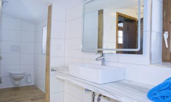 A modern bathroom with white tiles and a sink on a wooden shelf. In the background, a toilet and a mirror are visible.