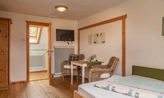 A cozy room with a wooden floor, a bed, a table, and two chairs. The room has daylight through a window and is located in a quiet ambiance.