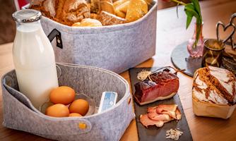 A breakfast table with various baked goods and a container of milk and eggs. On a black plate, ham and bread are arranged.