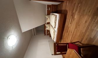 A cozy bedroom with two beds, wooden floors, and a table. The walls are in neutral colors and there is a large window area.