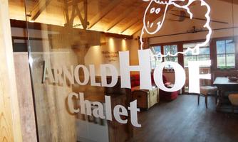A cozy chalet with a warm, inviting interior. A transparent sign displays the name "ARNOLD HOF Chalet" and a sheep logo.
