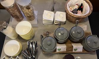 A breakfast buffet with bowls for muesli, yogurt, and toppings. There are also plates, spoons, and a small basket with sweets.