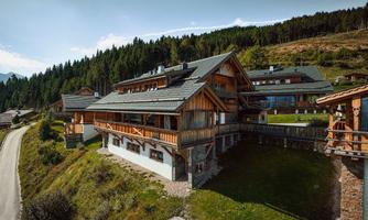 A modern wooden house in the mountains with large balconies. Surrounded by green hills and trees.